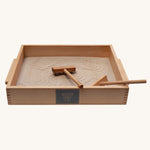 Wooden Story Montessori Sand Tray and Accessories shown on a plain background. The sand is in the tray with the tools. The tray is placed at an angle and the printed logo can be seen on the side. 