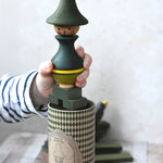 A close up of a child's hand holding the Wooden Story green stick figure stacking toy. The patterned gift box can be seen underneath. 