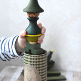 A close up of a child's hand holding the Wooden Story green stick figure stacking toy. The patterned gift box can be seen underneath. 