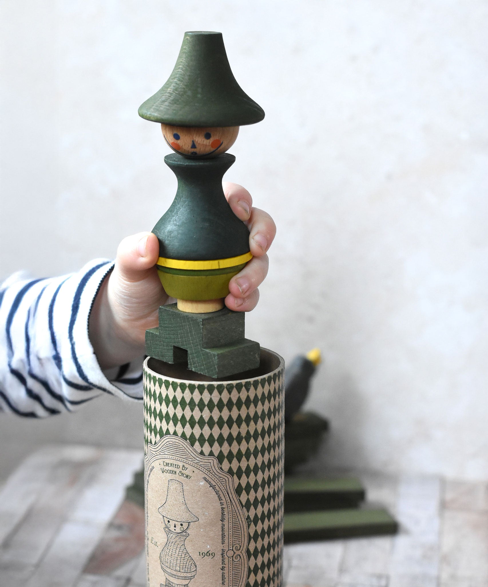 A close up of a child's hand holding the Wooden Story green stick figure stacking toy. The patterned gift box can be seen underneath. 