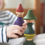 A Wooden Story green stick figure stacking toy on a wooden surface. A child can be seen in the background holding onto the purple stick figure. 