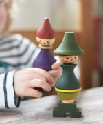A Wooden Story green stick figure stacking toy on a wooden surface. A child can be seen in the background holding onto the purple stick figure. 