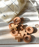 The wooden X and O pieces from the Wooden Story tic tac toe game laid out on a white wooden surface, the cotton drawstring storage bag can be seen in the background.