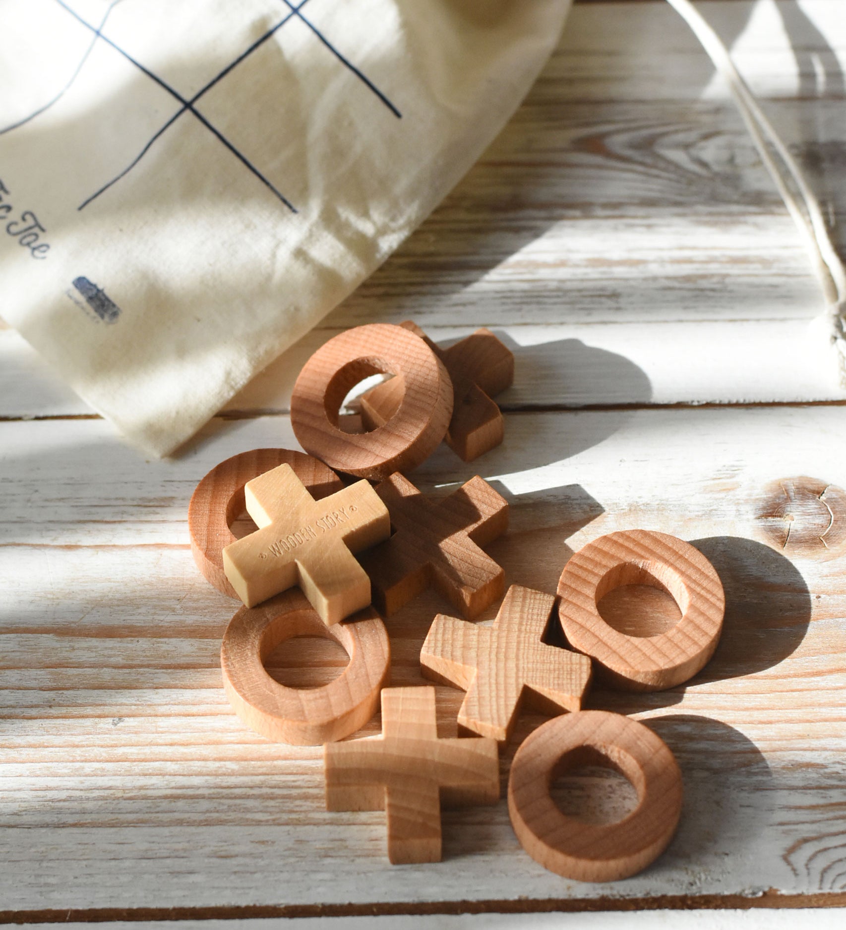 The wooden X and O pieces from the Wooden Story tic tac toe game laid out on a white wooden surface, the cotton drawstring storage bag can be seen in the background.
