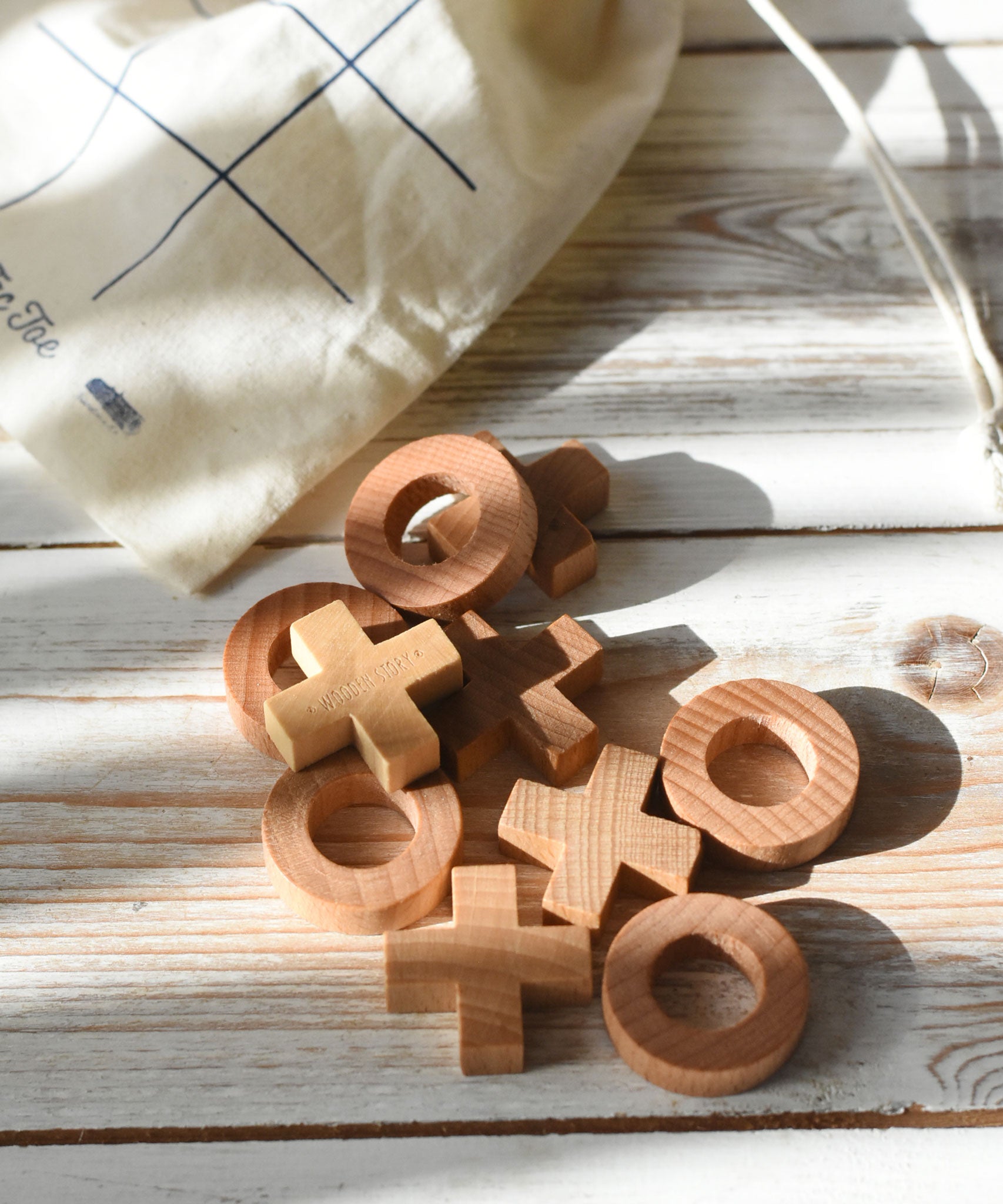 The wooden X and O pieces from the Wooden Story tic tac toe game laid out on a white wooden surface, the cotton drawstring storage bag can be seen in the background.