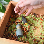 A Wooden Story Play Tray filled with coloured dried rice and chick peas. A child's hand can be seen placing more rice in the tray n top of a blue coloured Grimm's friends and a Ostheimer black bird.