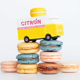 Picture of the Citron Candylab Candyvan on top of a pile of macarons. It is a yellow and white van with a yellow Lemon Macaron on the roof.