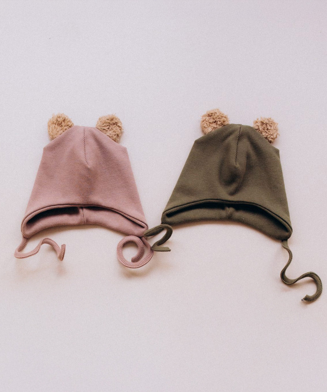 Wooly Organic hat with teddy ears in pale blush / savannah next to the tea leaf green coloured hat both available at Babipur. A GOTS organic hat in a soft ribbed style. Features ties and cute fluffy brown teddy bear ears
