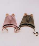 Wooly Organic hat with teddy ears in pale blush / savannah next to the tea leaf green coloured hat both available at Babipur. A GOTS organic hat in a soft ribbed style. Features ties and cute fluffy brown teddy bear ears