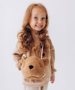 A child wearing the Wooly Organic Kids' Teddy Bear Crossbody Bag. The classic Wooly Organic bear turned into a cute wearable bag for children. A range of Wooly Organic bears and clothing are available at Babipur.