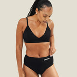 Lifestyle image of a model wearing the WUKA Stretch™ Seamless Midi Brief period underwear with a matching black bra, showing the full outfit and how the period pants sit on the body.