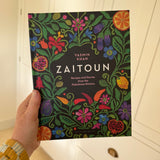 A hand holding the Zaitoun: Recipes and Stories from the Palestinian Kitchen hardback cookbook. The cover is black with mock-embroidered flowers and fruits. 
