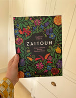 A hand holding the Zaitoun: Recipes and Stories from the Palestinian Kitchen hardback cookbook. The cover is black with mock-embroidered flowers and fruits. 