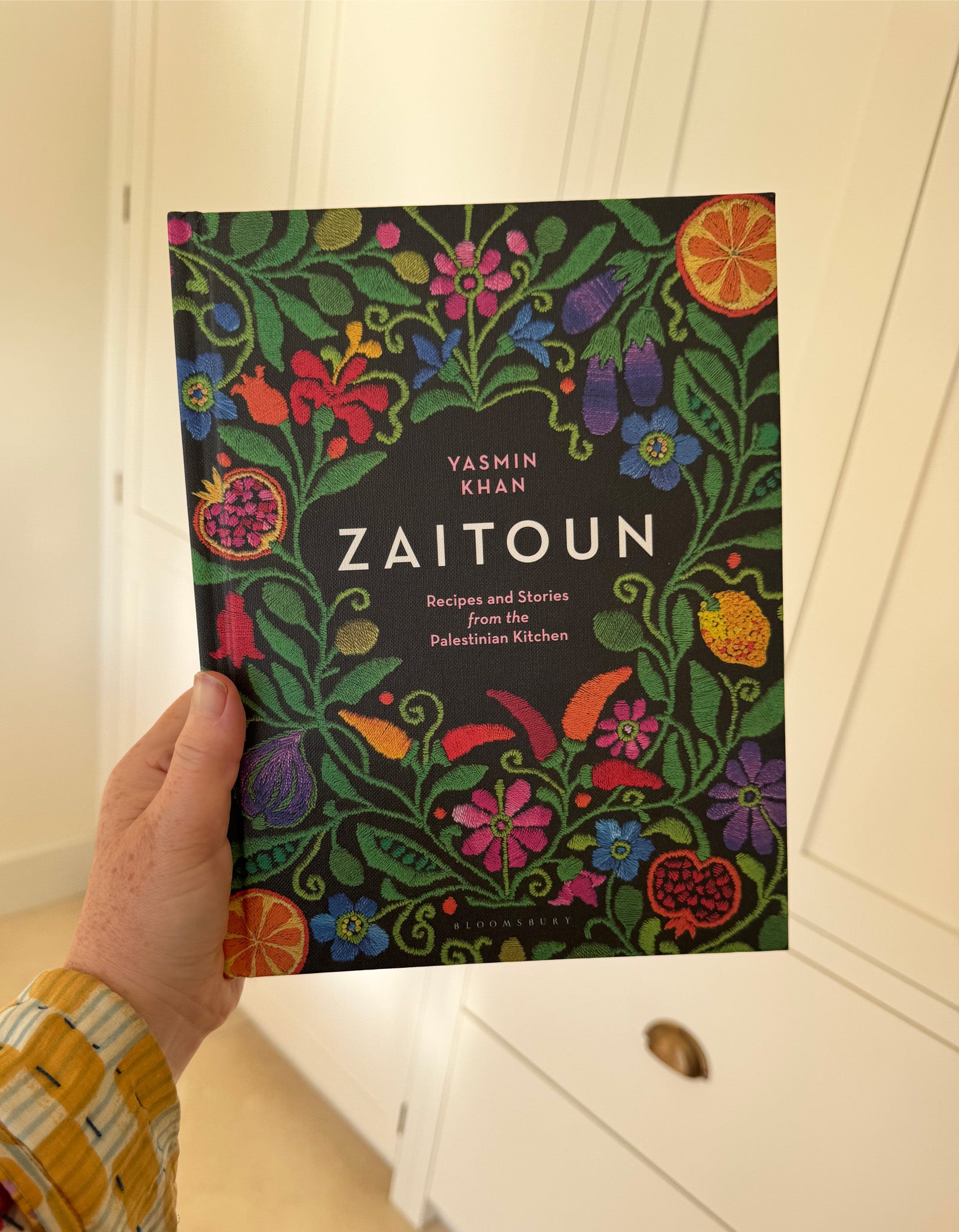 A hand holding the Zaitoun: Recipes and Stories from the Palestinian Kitchen hardback cookbook. The cover is black with mock-embroidered flowers and fruits. 