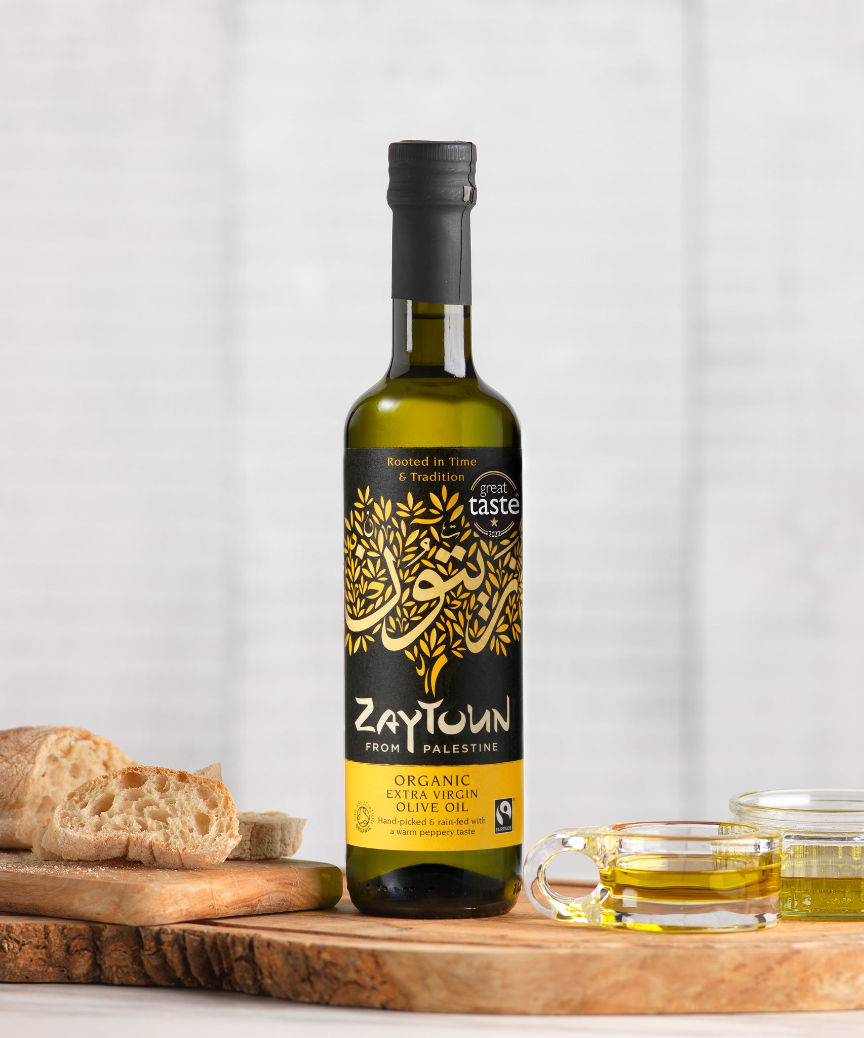 A 250ml bottle of Zaytoun Organic Extra Virgin Olive Oil on a wooden board next to bread and glass dishes containing some olive oil. 